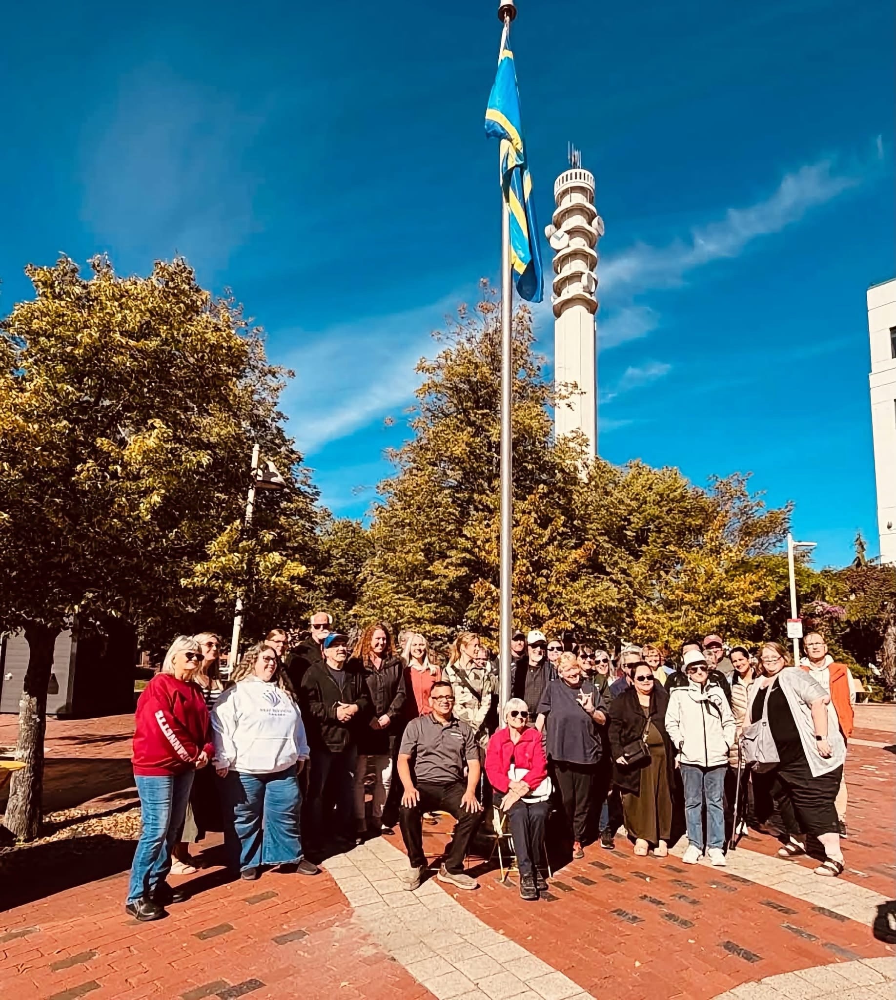 Community members gathered outdoors by a flagpole under a bright blue sky