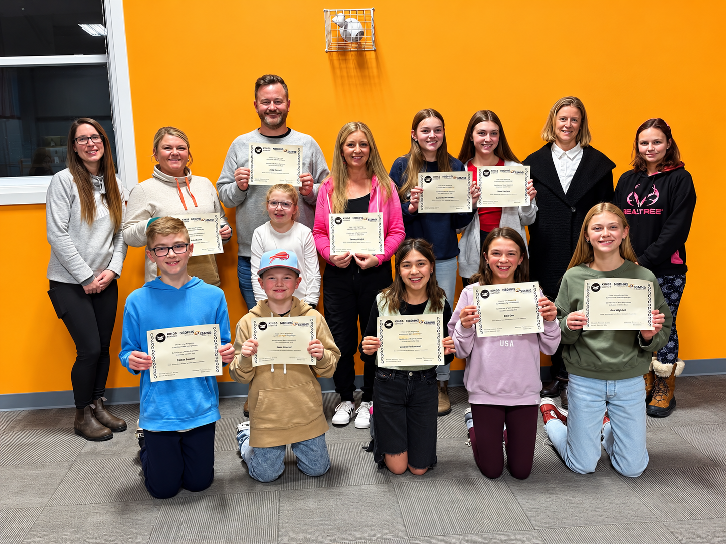 Group of ASL class graduates holding certificates in front of an orange wall